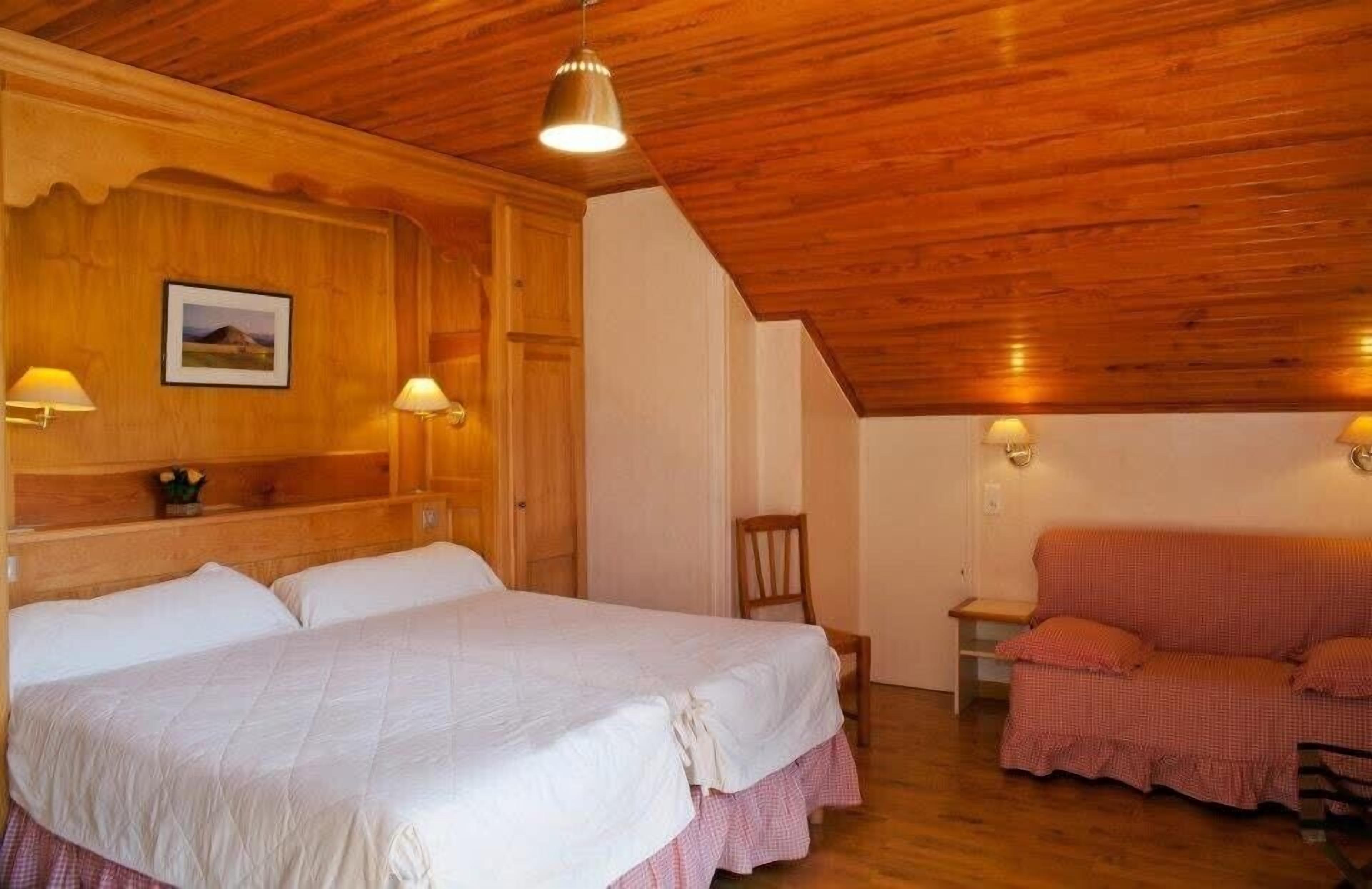 Une pièce avec un plafond en bois, un lit recouvert de linge de lit blanc, un canapé à carreaux rouges et deux lampes fixées au mur.