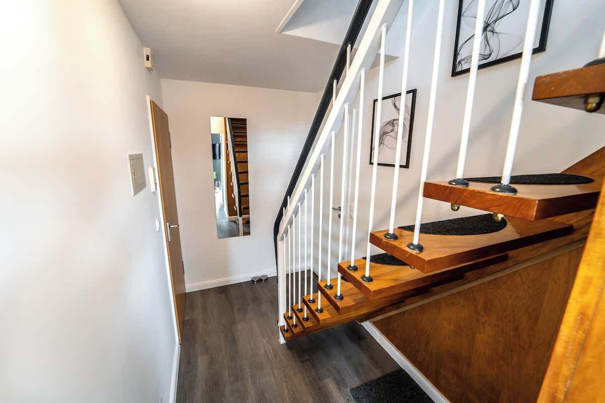 Una escalera moderna con peldaños de madera y barandillas blancas, que conduce a un descansillo con un espejo y un cuadro enmarcado.