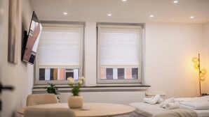 Moderni hotellihuone, jossa on kaksi suurta ikkunaa, pyöreä pöytä maljakolla, siististi taitelluilla pyyhkeillä varustettu sänky ja lattiavalaisin.