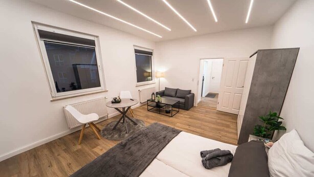 Een moderne, goed verlichte kamer met een bed, een slaapbank, een salontafel en een klein tafeltje met een stoel. Er zijn ramen en een deur zichtbaar.