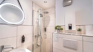 Un baño moderno con ducha envuelta en vidrio, espejo redondo, lavamanos blanco y una pequeña planta en maceta.