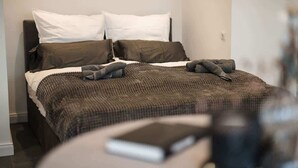 A neatly made bed with a dark brown blanket and pillows, a bedside table with a book, and a plain wall with a light switch. - Exklusive 1-Zimmer Stadtwohnung in Bremen (Bremen)