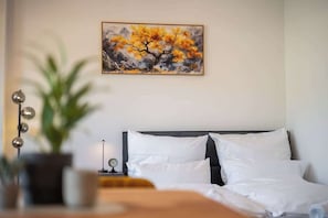 A hotel room with a bed, white pillows, a nightstand with a clock, and a painting of a tree on the wall. - Steintorperle – 2 Zimmer im Herzen vom Viertel (Bremen)