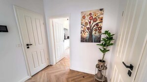 Un couloir avec un parquet en chevrons, une porte blanche, un tableau représentant un arbre et une plante en pot.