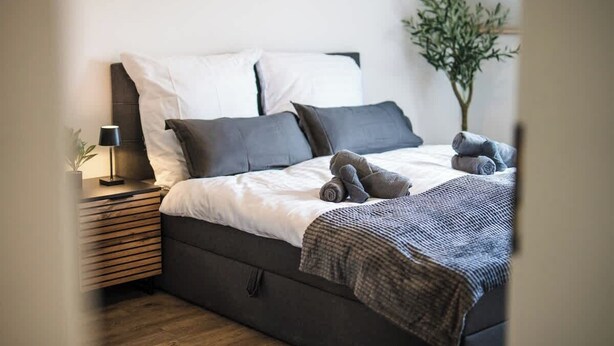 Ett modernt sovrum med en säng, kuddar, ett sängbord med en lampa och en liten växt.