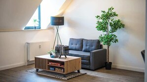 Ett modernt vardagsrum med ett sluttande tak, en mörkgrå soffa, ett soffbord i trä, en golvlampa och en krukväxt.