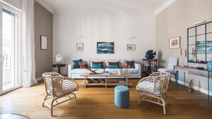 Een ruime woonkamer met een witte bank, twee rieten fauteuils, een salontafel en een blauwe poef. Er hangen kunstwerken in lijsten aan de muren en er is een groot raam met gordijnen.