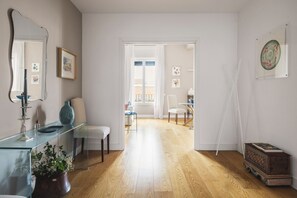Ein modernes Wohnzimmer mit einem Glastisch, einem weißen Sofa und Holzboden. Eine Tür führt in einen anderen Raum mit einem Balkon.