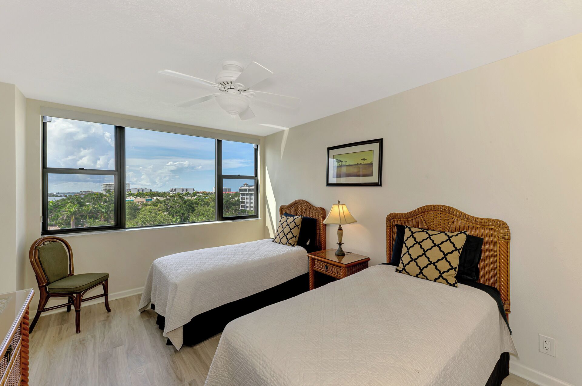 Ein Hotelzimmer mit zwei Betten, einem Sessel, einem Nachttisch mit Lampe, einem Deckenventilator und einem großen Fenster mit Blick auf die Stadt.