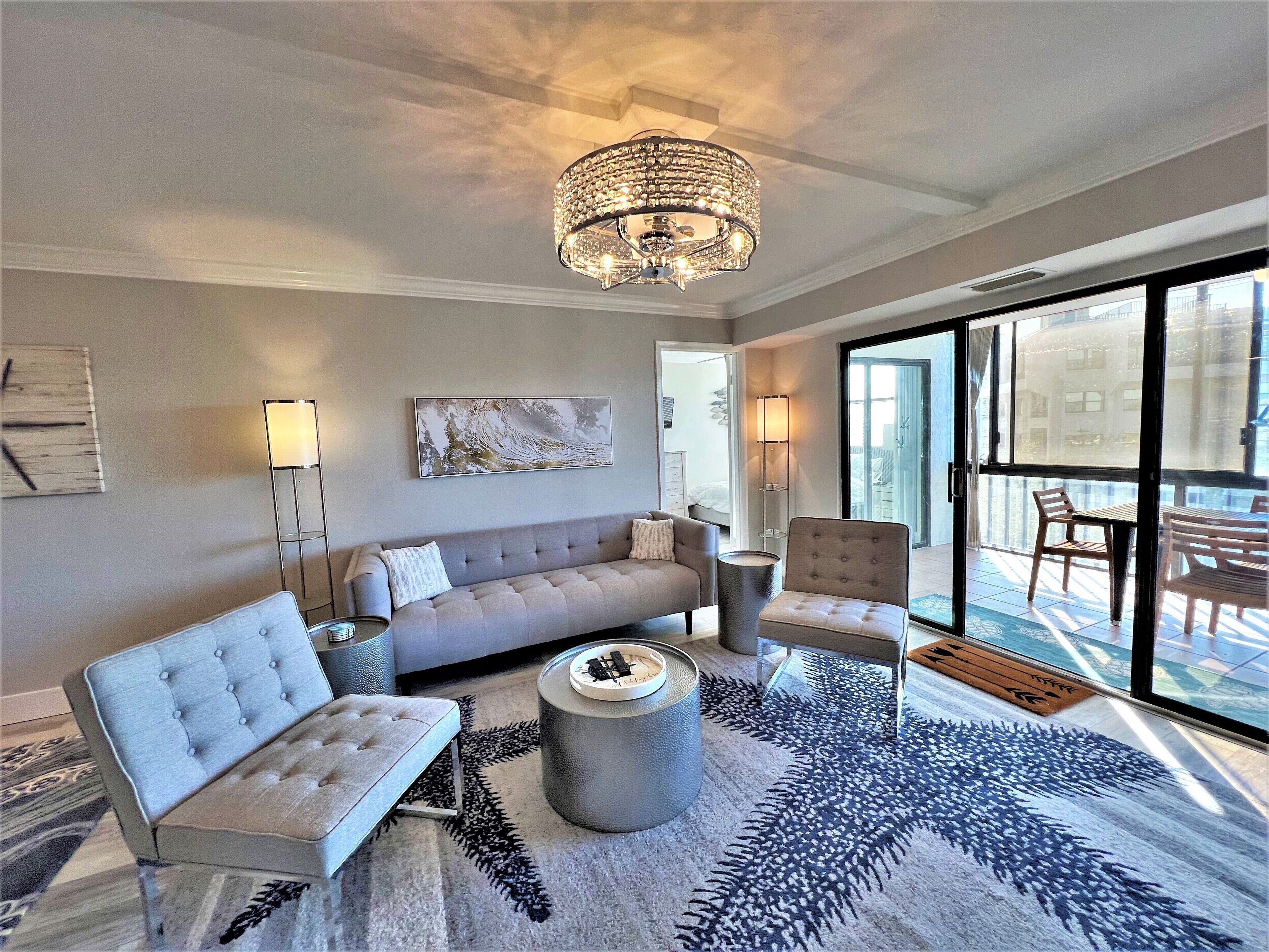 A modern living room with a grey sofa, a round ottoman, and a chandelier. There is a large window with a view of the outside.