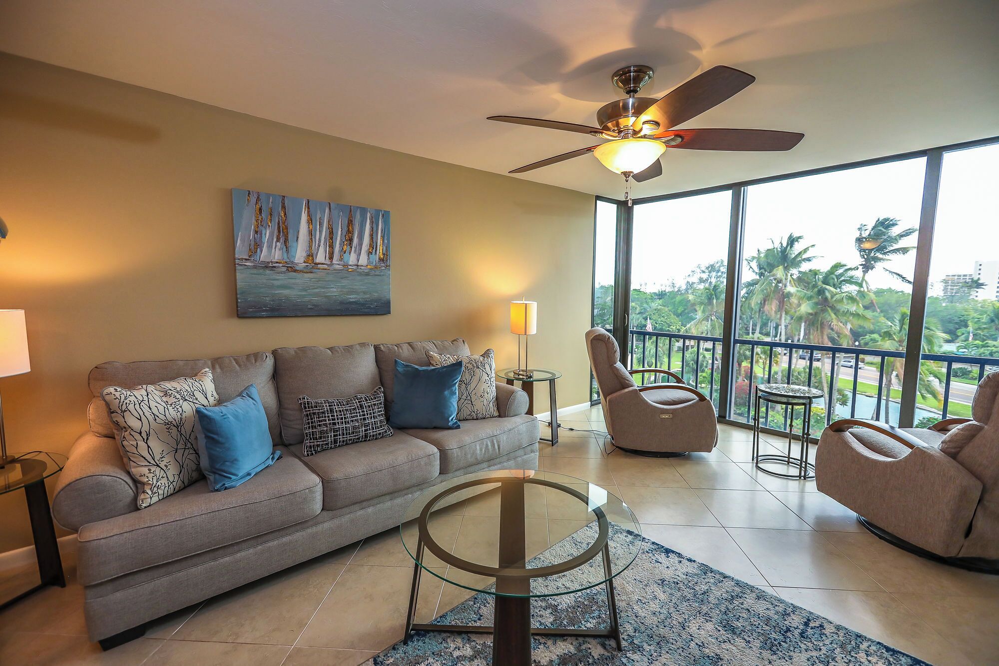 A living room with a sofa, armchairs, a glass coffee table, a painting, a ceiling fan, and a view of palm trees through large windows.