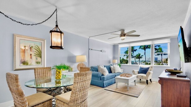 A modern living room with a glass dining table, wicker chairs, a blue sofa, and a wooden cabinet. - Harbor Towers 424 - Tropical Sands Accommodations (Siesta Key)