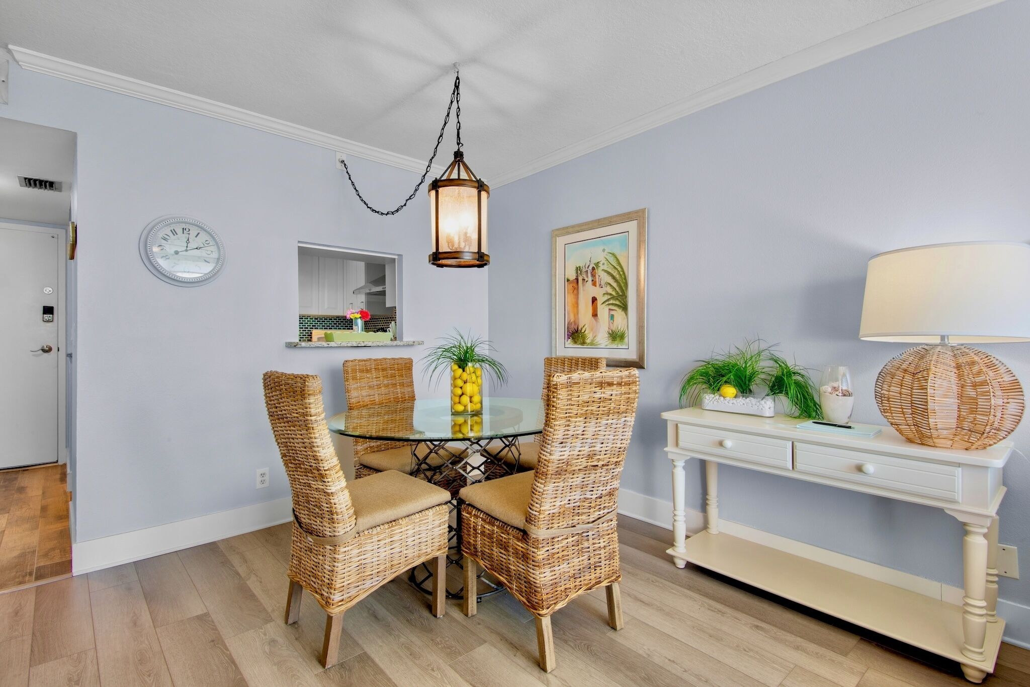Een eethoek met rieten stoelen, een glazen tafel, een hanglamp, een wandklok, een ingelijste foto en een console tafel met decoratieve objecten.