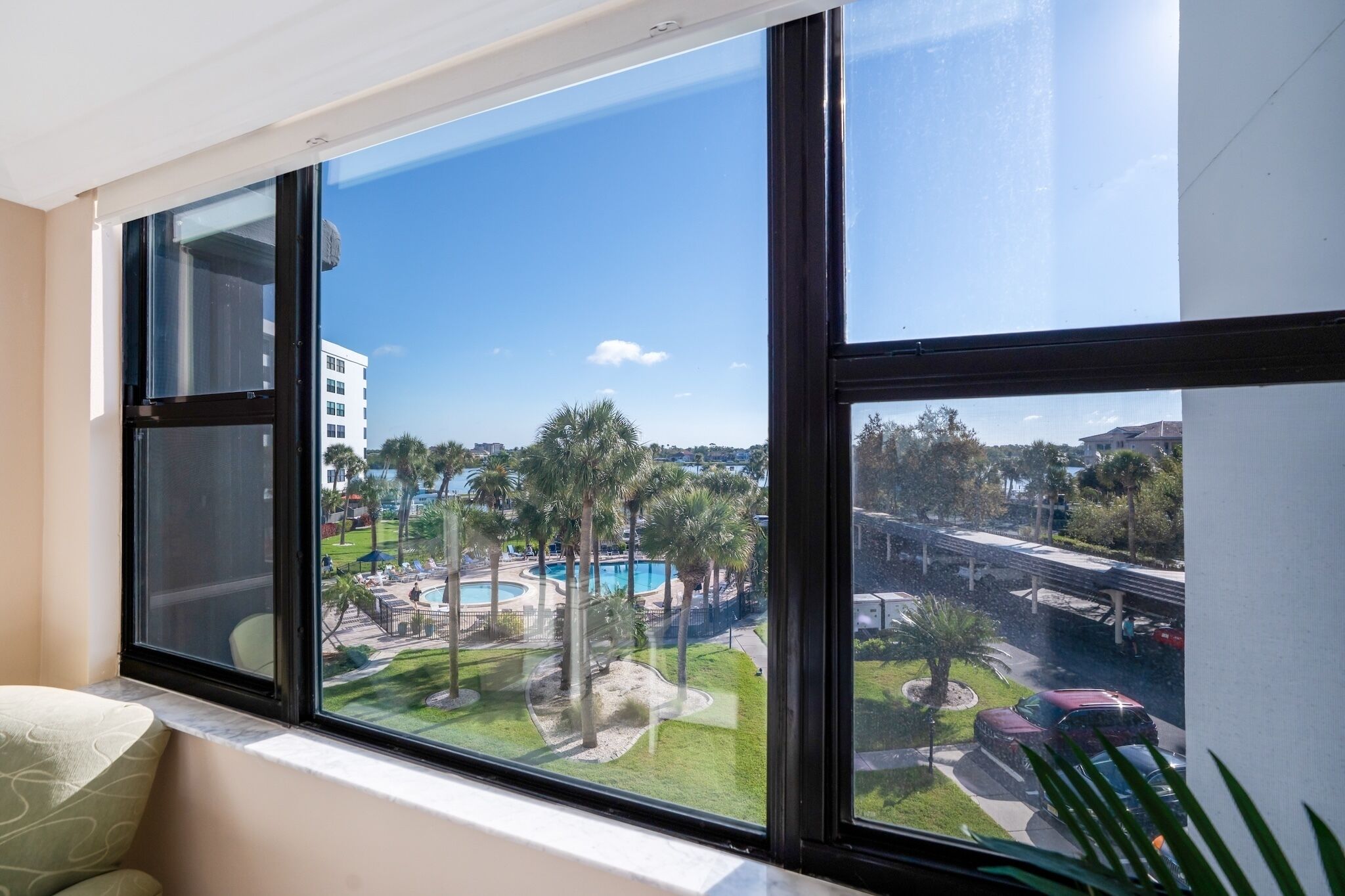 Ein Zimmer mit großem Fenster mit Blick auf einen Poolbereich mit Palmen und einem Gebäude im Hintergrund.