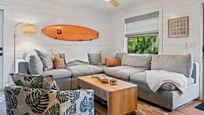 A modern living room with a sectional sofa, a wooden coffee table, a ceiling fan, and a surfboard decoration. - Luxury Family Getaway w/ Pool, Putting and Backyard Fun (Anna Maria)