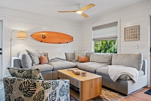 A modern living room with a sectional sofa, a wooden coffee table, a ceiling fan, and a surfboard decoration. - Luxury Family Getaway w/ Pool, Putting and Backyard Fun (Anna Maria)
