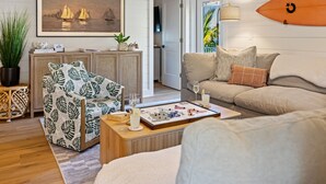 A living room with a sofa, a patterned armchair, a coffee table with a board game, a surfboard on the wall, and a painting of sailboats. - Luxury Family Getaway w/ Pool, Putting and Backyard Fun (Anna Maria)