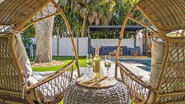 A wicker lounge chair with a bottle and glasses on a woven table, a pool, and a covered seating area in a tropical setting. - Luxury Family Getaway w/ Pool, Putting and Backyard Fun (Anna Maria)