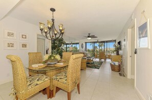 A spacious living area with a dining table surrounded by wicker chairs, a chandelier, and a large window offering a view of palm trees. - Harbor Towers 326 - Tropical Sands Accommodations (Siesta Key)