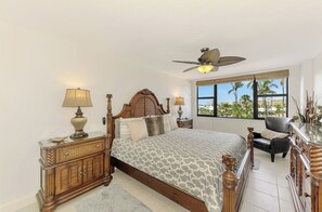 A bedroom with a bed, two nightstands, a ceiling fan, a window with a view of palm trees, and a chair. - Harbor Towers 326 - Tropical Sands Accommodations (Siesta Key)