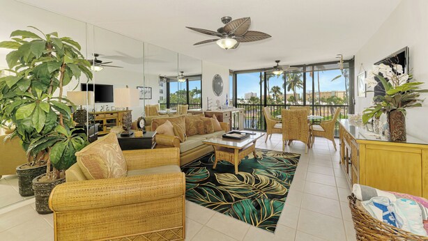 A spacious living room with a large sofa, a coffee table, and a dining area with chairs. There is a ceiling fan, a TV, and a large window offering a view of palm trees. - Harbor Towers 326 - Tropical Sands Accommodations (Siesta Key)
