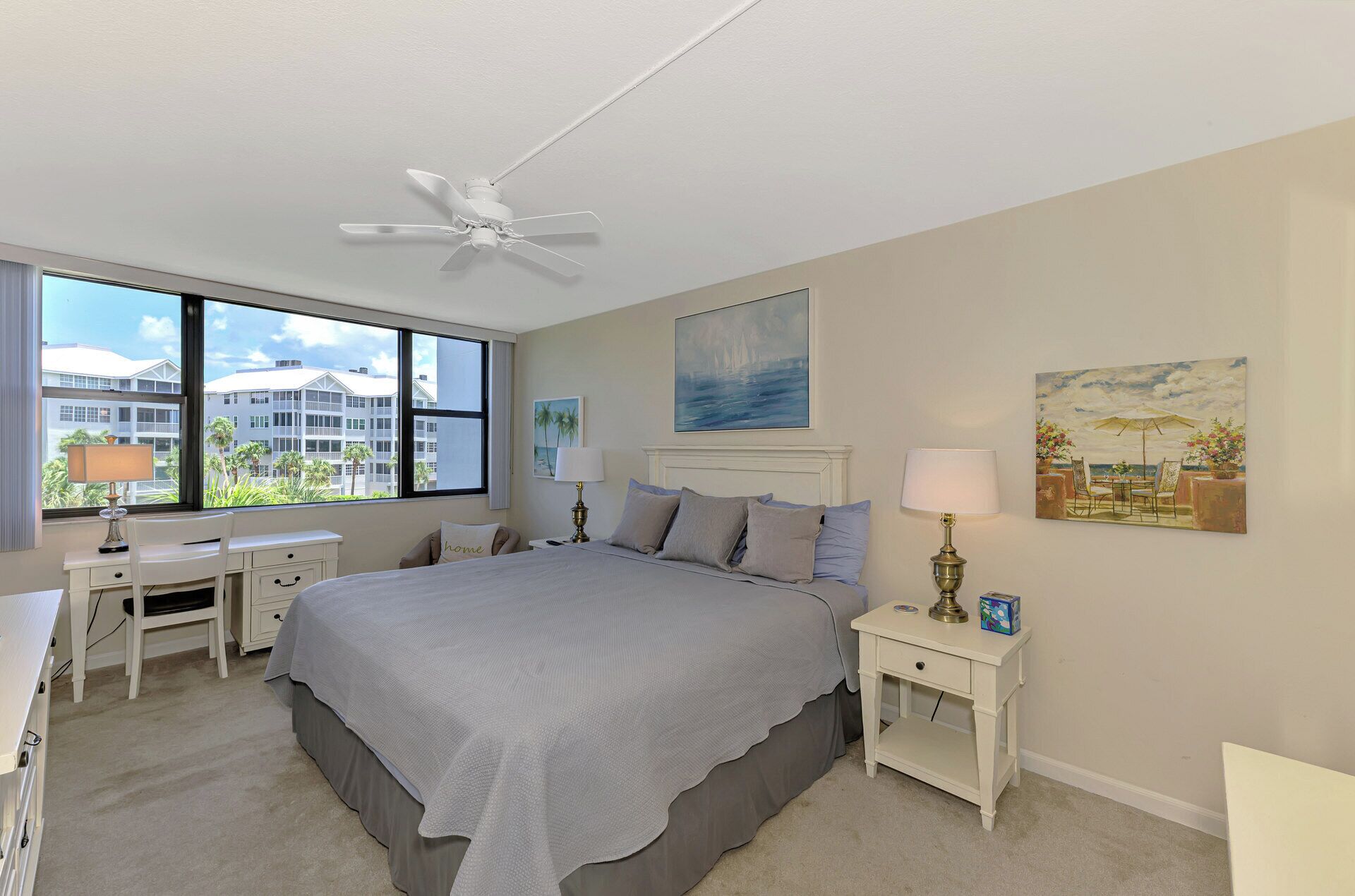 A bedroom with a large bed, a ceiling fan, a desk, and a window with a view of buildings.