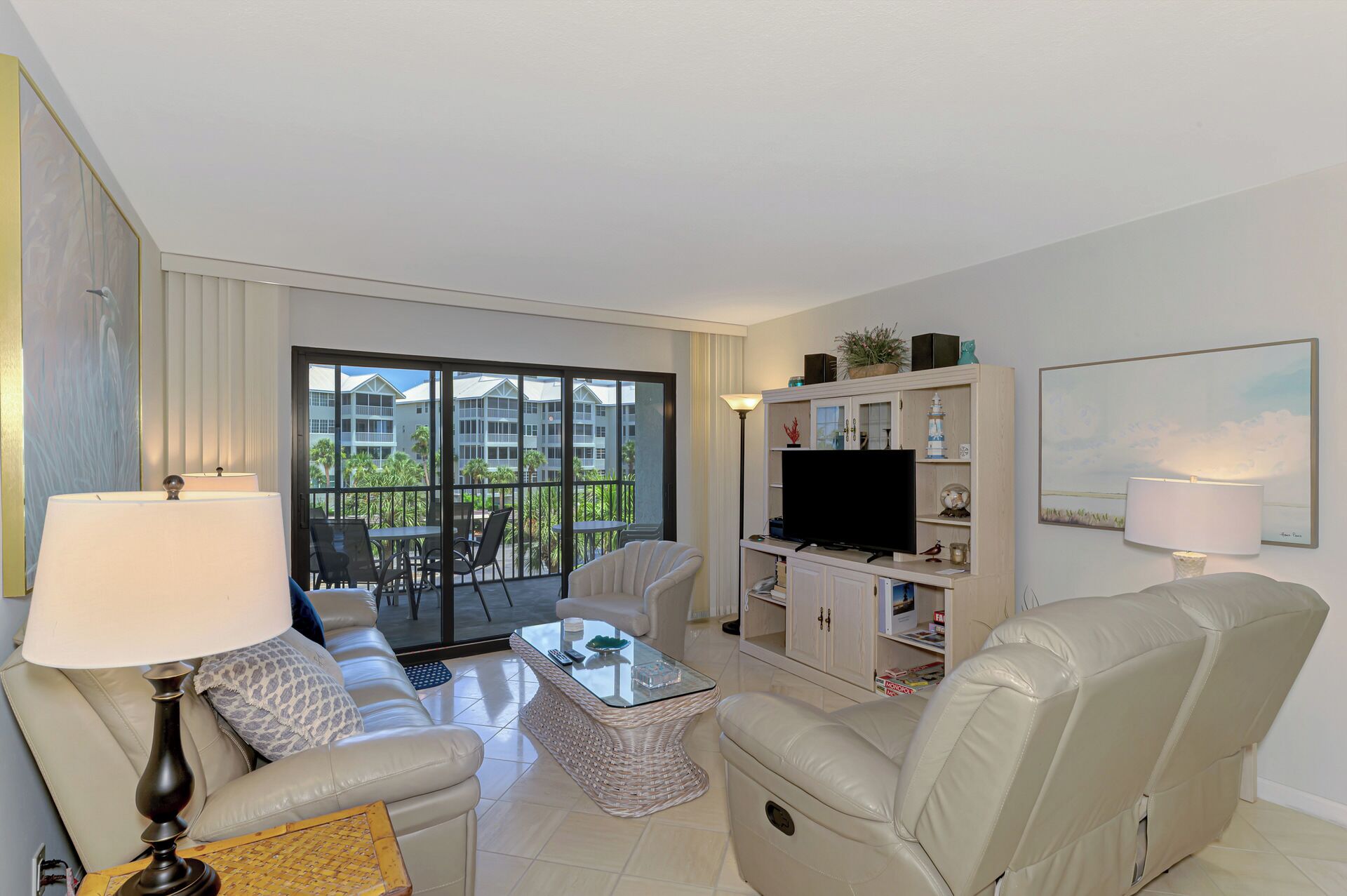 Ein modernes Wohnzimmer mit einer Couch, Sesseln, einem Couchtisch und einem Fernseher. Ein Balkon bietet Blick auf Gebäude und Palmen.