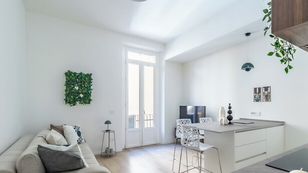 Een moderne keuken met witte kastjes, een eethoek met witte stoelen, en een woonkamer met een bank en decoratieve kussens.
