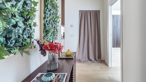 Een hal met een houten vloer, een spiegel, een aan de muur bevestigde plant, een vaas met bloemen en een deur met gordijn.