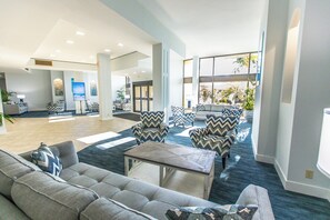 A spacious lobby with a modern design, featuring a large seating area with patterned chairs and sofas, a reception desk, and a view of the outdoors through large windows. - Harbor Towers 303 - Tropical Sands Accommodations (Siesta Key)