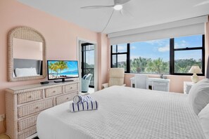Een hotelkamer met een bed, een televisie, een bureau en uitzicht op palmbomen en een strand.