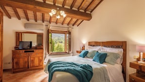 A bedroom with a wooden ceiling, a large bed with multiple pillows, a nightstand with a lamp, a dresser with a TV, and a window with a view of greenery. - CASA VECCIANO essence of Tuscany (Toscana)