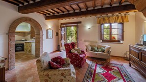 A rustic living room with exposed wooden beams, a brick archway leading to a kitchen, and a cozy seating area with patterned sofas. - CASA VECCIANO essence of Tuscany (Toscana)