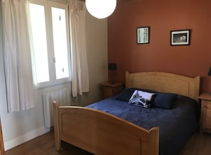 Ein Schlafzimmer mit einem hölzernen Bett, einem Nachttisch, einem Fenster mit Vorhängen und zwei an der Wand hängenden Bildern.