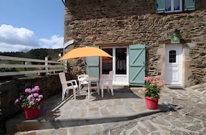 A stone building with a patio area featuring outdoor seating, a potted plant, and a window with shutters.