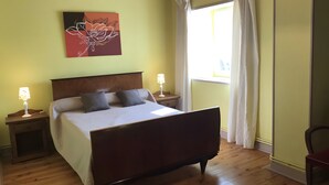 A bedroom with a wooden bed, two bedside tables with lamps, a window with curtains, and a wall art piece.