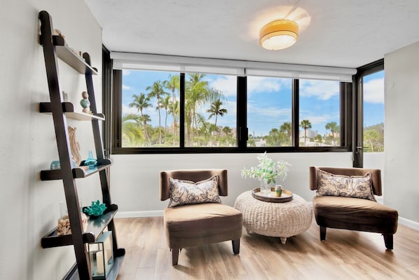 A room with two brown armchairs, a small round table, and a black shelving unit against a white wall. A large window offers a view of palm trees and buildings. - Harbor Towers 221 - Tropical Sands Accommodations (Siesta Key)