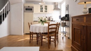 Un coin repas avec une table dressée pour quatre personnes, des chaises en bois, une nappe blanche et un vase de fleurs. On aperçoit une cuisine en arrière-plan, avec des placards et des appareils électroménagers.
