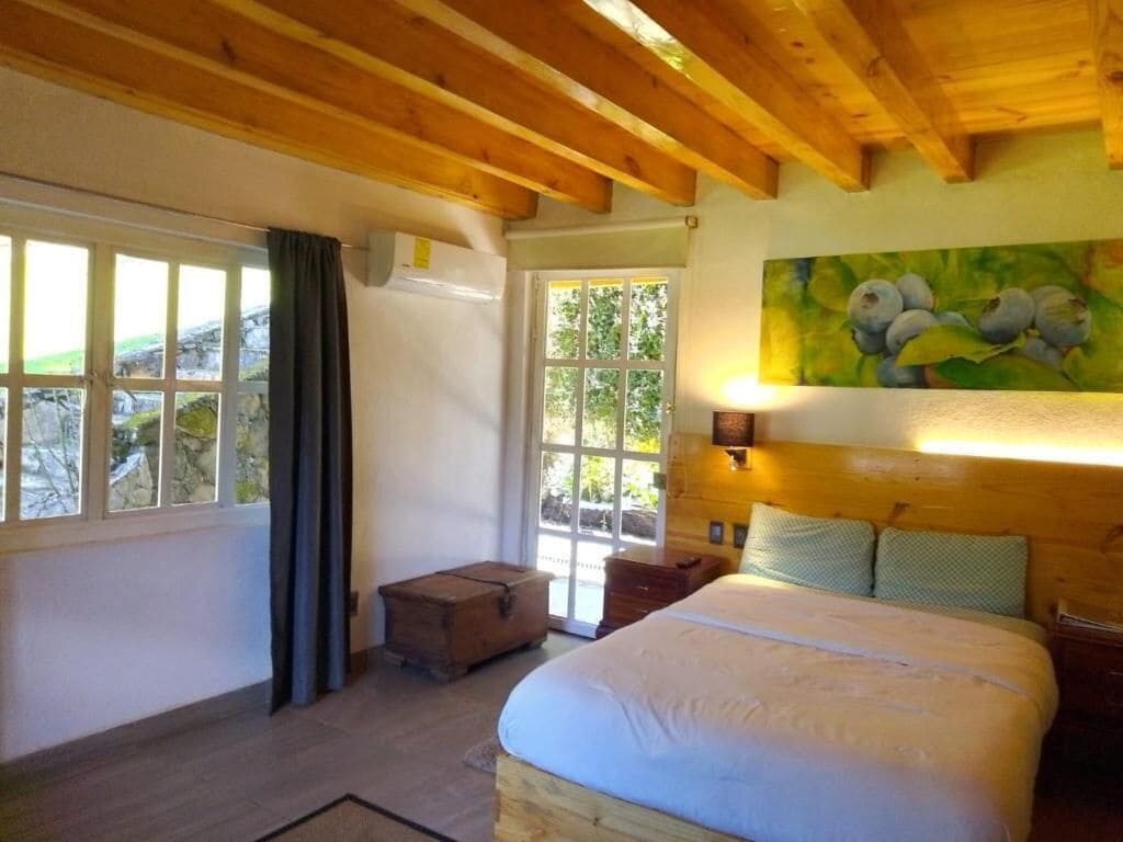 Una camera da letto con soffitto in legno, un letto, un comodino, una finestra con tende e un quadro appeso al muro.