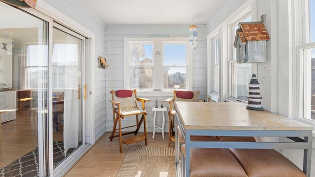 Ein heller Raum mit einem Holztisch und Stühlen, einem kleinen weißen Tisch und Blick ins Freie durch große Fenster.