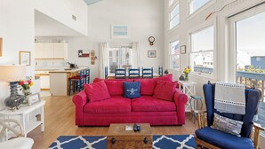 Ein großzügiges Wohnzimmer mit einem roten Sofa, einem Holztisch und einem blauen Sessel. Im Hintergrund ist eine Küche mit Essbereich zu sehen.