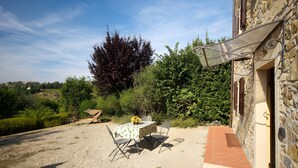 Eine Terrasse im Freien mit Tisch und Stühlen, umgeben von Pflanzen und einem Steinhaus.