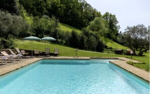 Una piscina rectangular con una superficie de agua azul cristalina, rodeada por una solana de hormigón. Hay sillones de descanso y dos sombrillas verdes en la solana. La piscina tiene como fondo árboles verdes frondosos y una colina cubierta de hierba.