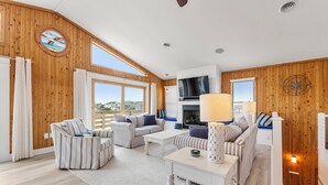 A spacious living room with a high ceiling, wooden walls, and large windows. There is a fireplace, a television, and a dining area with a table and chairs. - Tails in the Sand: Private Heated Pool | Hot Tub | Dogs OK (Corolla)