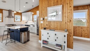 A modern kitchen with wooden paneling, white cabinets, stainless steel appliances, and a large island with bar stools. - Tails in the Sand: Private Heated Pool | Hot Tub | Dogs OK (Corolla)