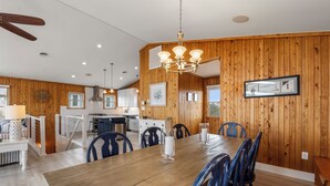 A dining area with a wooden table and chairs, a chandelier, and a kitchen in the background. - Tails in the Sand: Private Heated Pool | Hot Tub | Dogs OK (Corolla)
