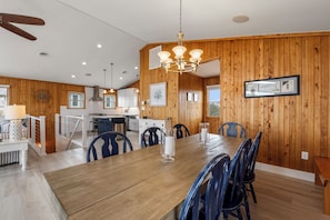A dining area with a wooden table and chairs, a chandelier, and a kitchen in the background. - Tails in the Sand: Private Heated Pool | Hot Tub | Dogs OK (Corolla)