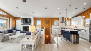 A spacious living area with a fireplace, a dining table, and a kitchen with white cabinets and stainless steel appliances. - Tails in the Sand: Private Heated Pool | Hot Tub | Dogs OK (Corolla)