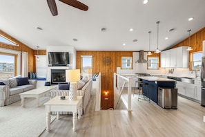 A spacious living area with a fireplace, a dining table, and a kitchen with white cabinets and stainless steel appliances. - Tails in the Sand: Private Heated Pool | Hot Tub | Dogs OK (Corolla)