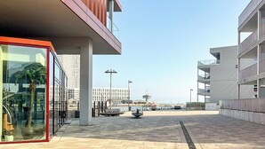 Un edificio moderno con fachada de vidrio, entrada enmarcada en rojo y un paseo empedrado que conduce al frente marítimo.